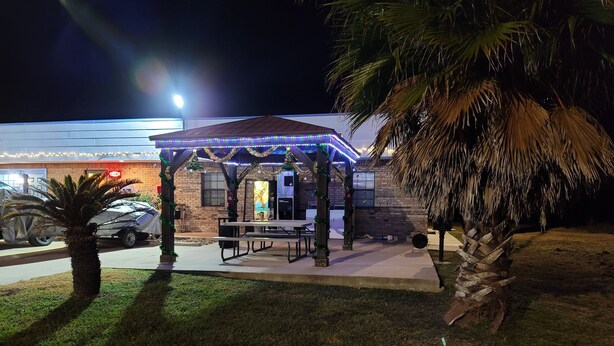 A covered outdoor seating area with a table and chairs, decorated with lights and greenery, in front of a building with a brick facade.