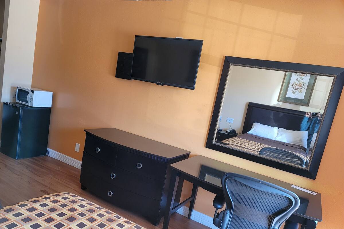 A hotel room with a bed, a dresser, a desk with a chair, a television mounted on the wall, and a large mirror reflecting the room.