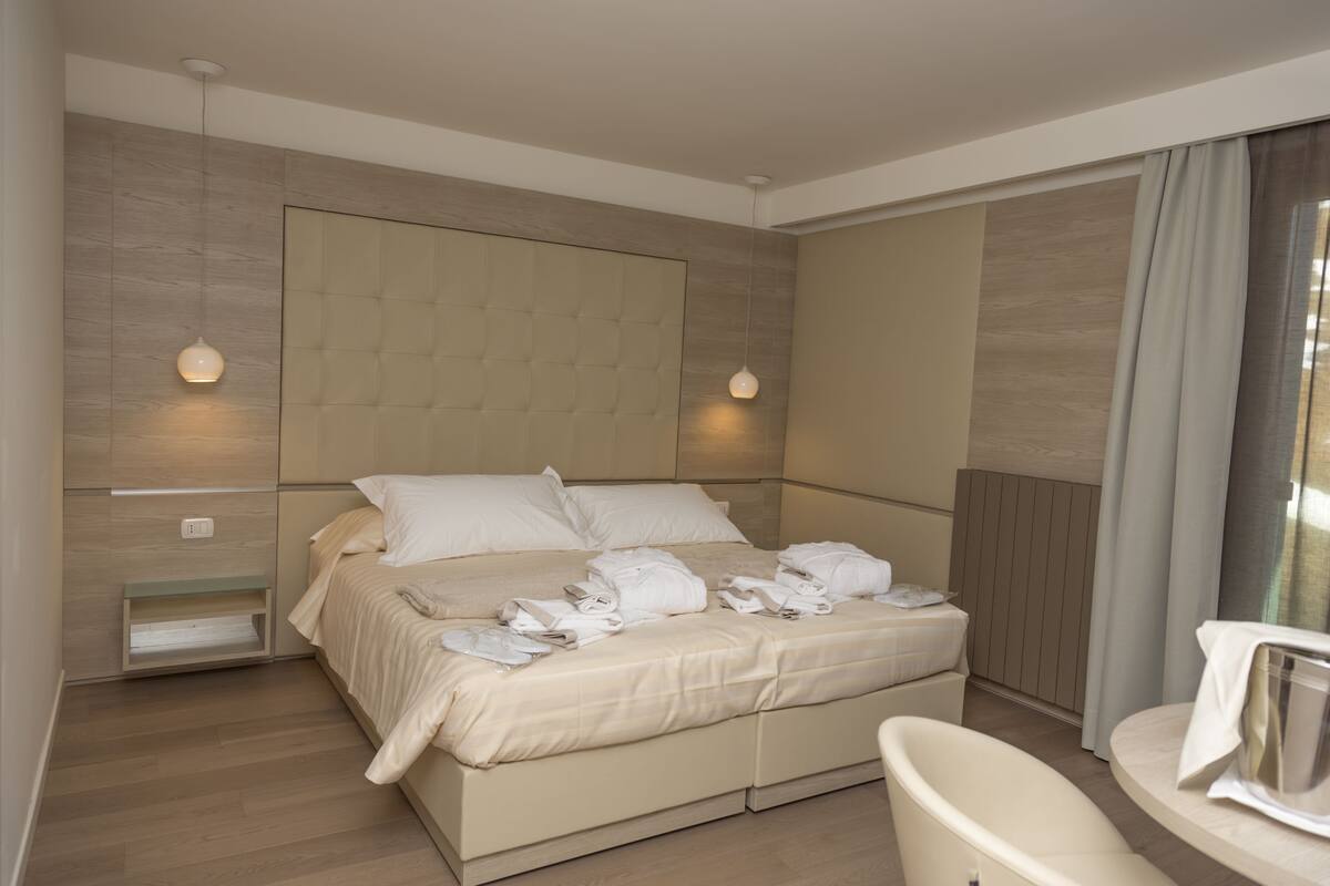 Habitación de hotel con una cama grande, mesitas de noche, cabecera incorporada y una pequeña zona de estar con una silla y una mesa.