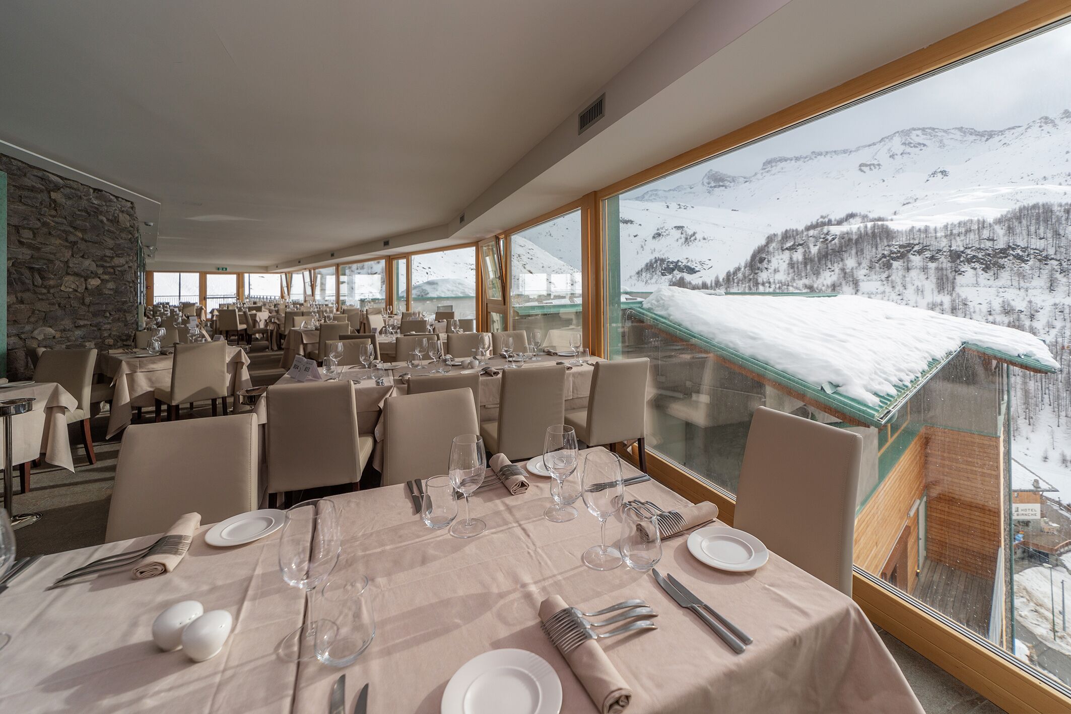 Un restaurant où les tables sont dressées pour le service, avec de grandes fenêtres donnant sur un paysage montagneux enneigé.