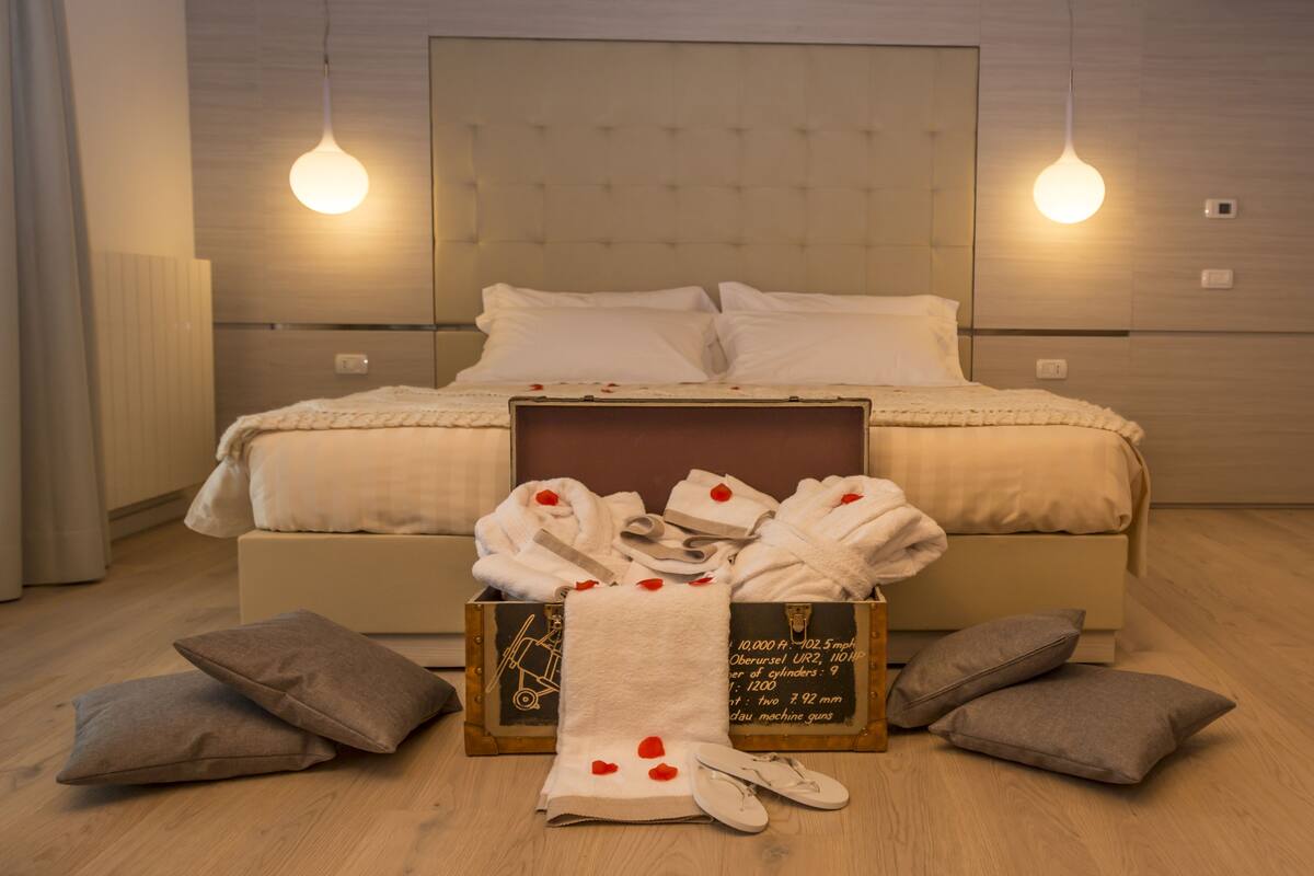Una cama bien hecha con ropa blanca, una mesita de noche con lámpara y un aparador de madera con toallas y pantuflas.