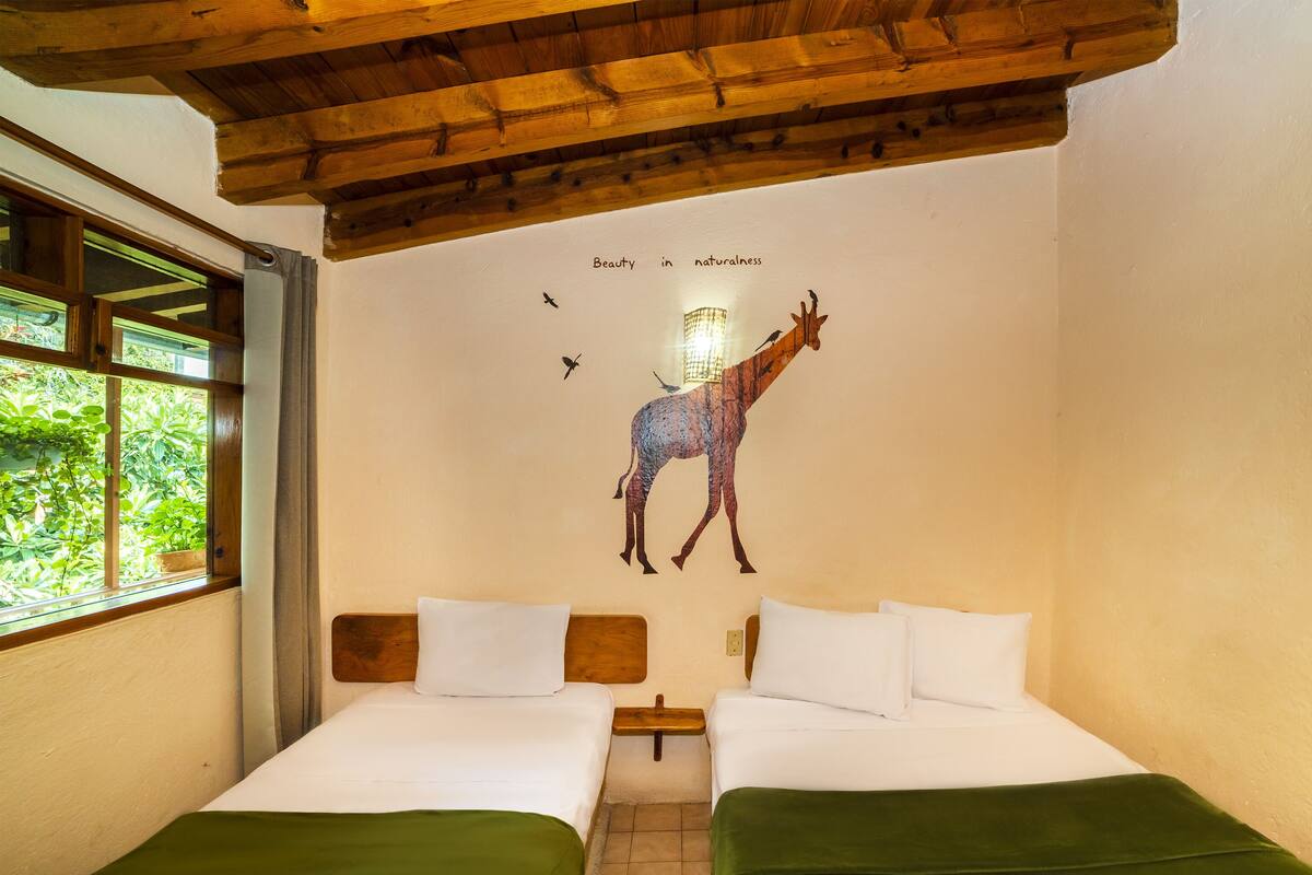 A room with two beds, a wooden ceiling, a wall mural of a giraffe, and a window with greenery outside.
