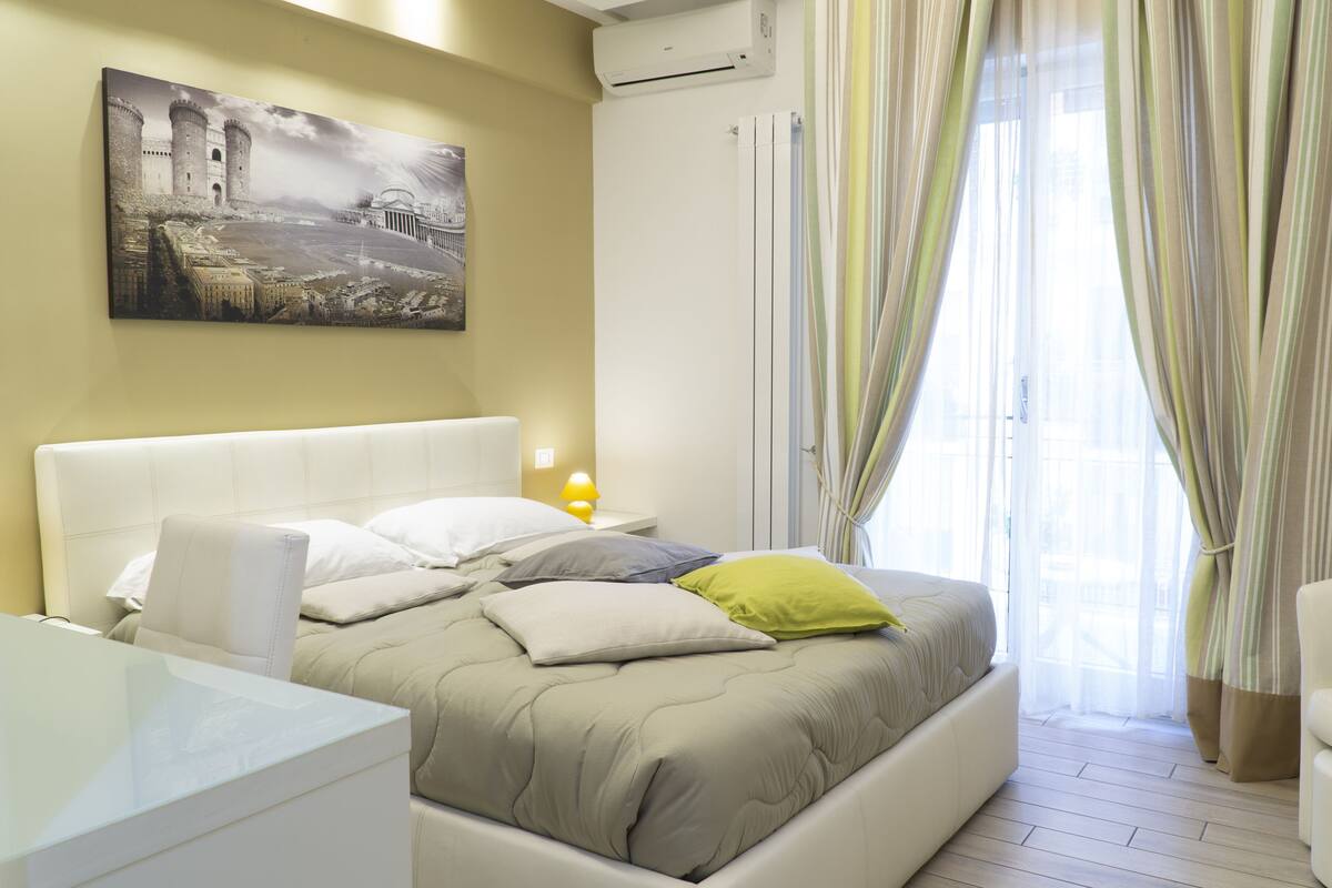 Un dormitorio con cama, almohadas, una mesita de noche, una lámpara y un cuadro en la pared.
