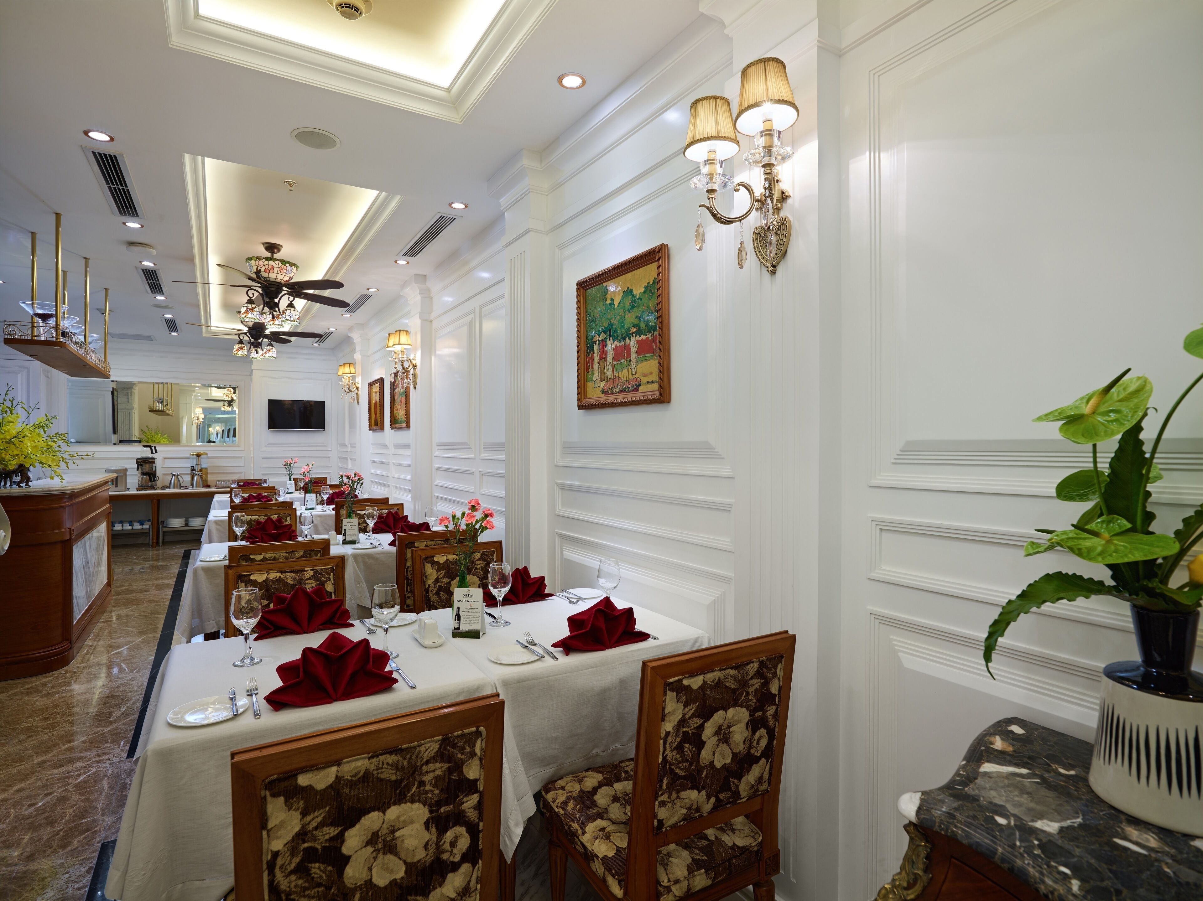 Un comedor formal con paredes blancas, candelabros y un ventilador de techo. Las mesas están dispuestas con servilletas rojas y cristalería.