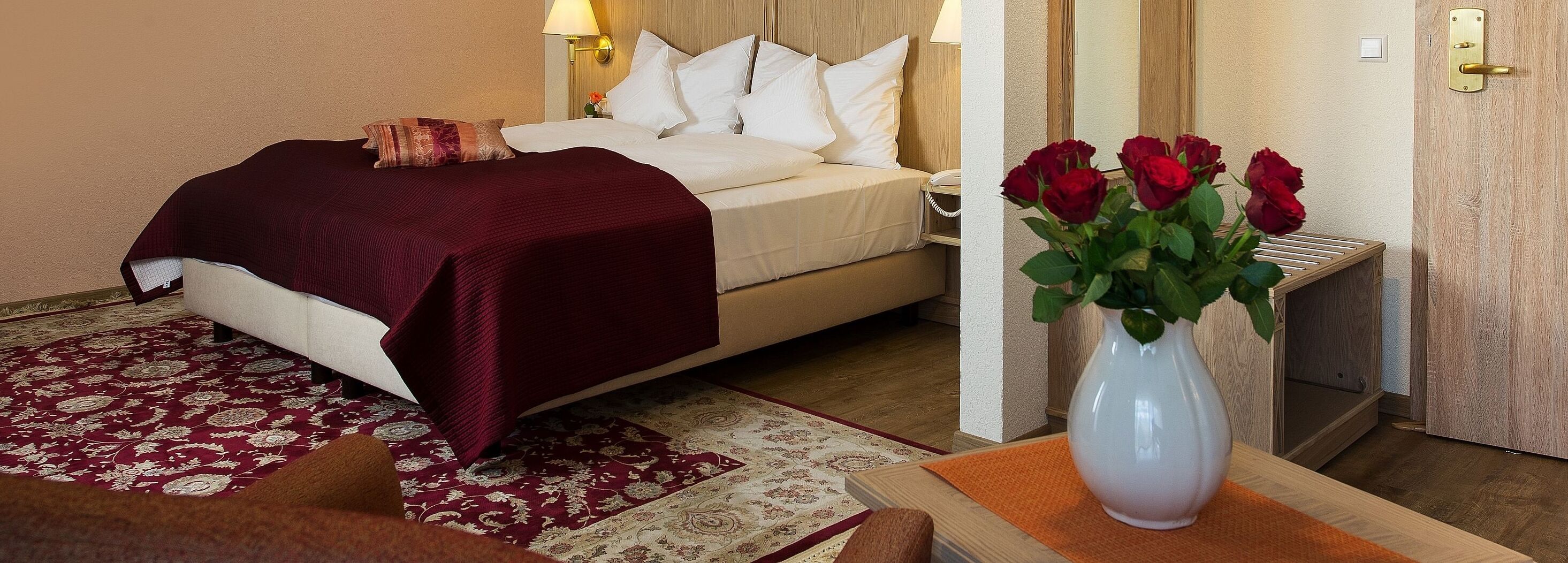 A hotel room with a bed, a nightstand, a lamp, a vase with red roses, and a wooden door.