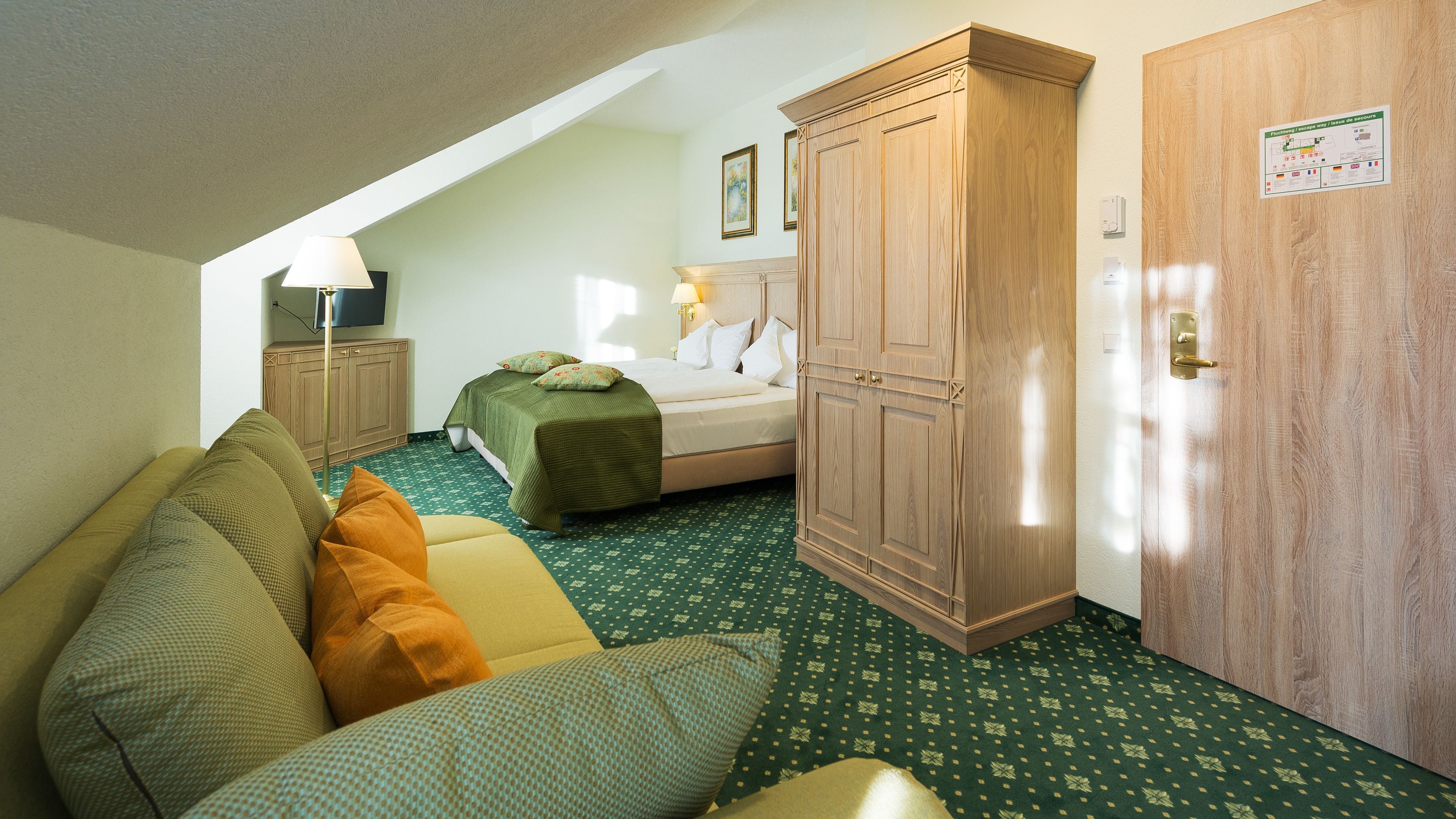 Habitació d'hotel amb un llit, un sofà verd amb dibuixos, un armari de fusta i una petita taula de nit amb una làmpada.