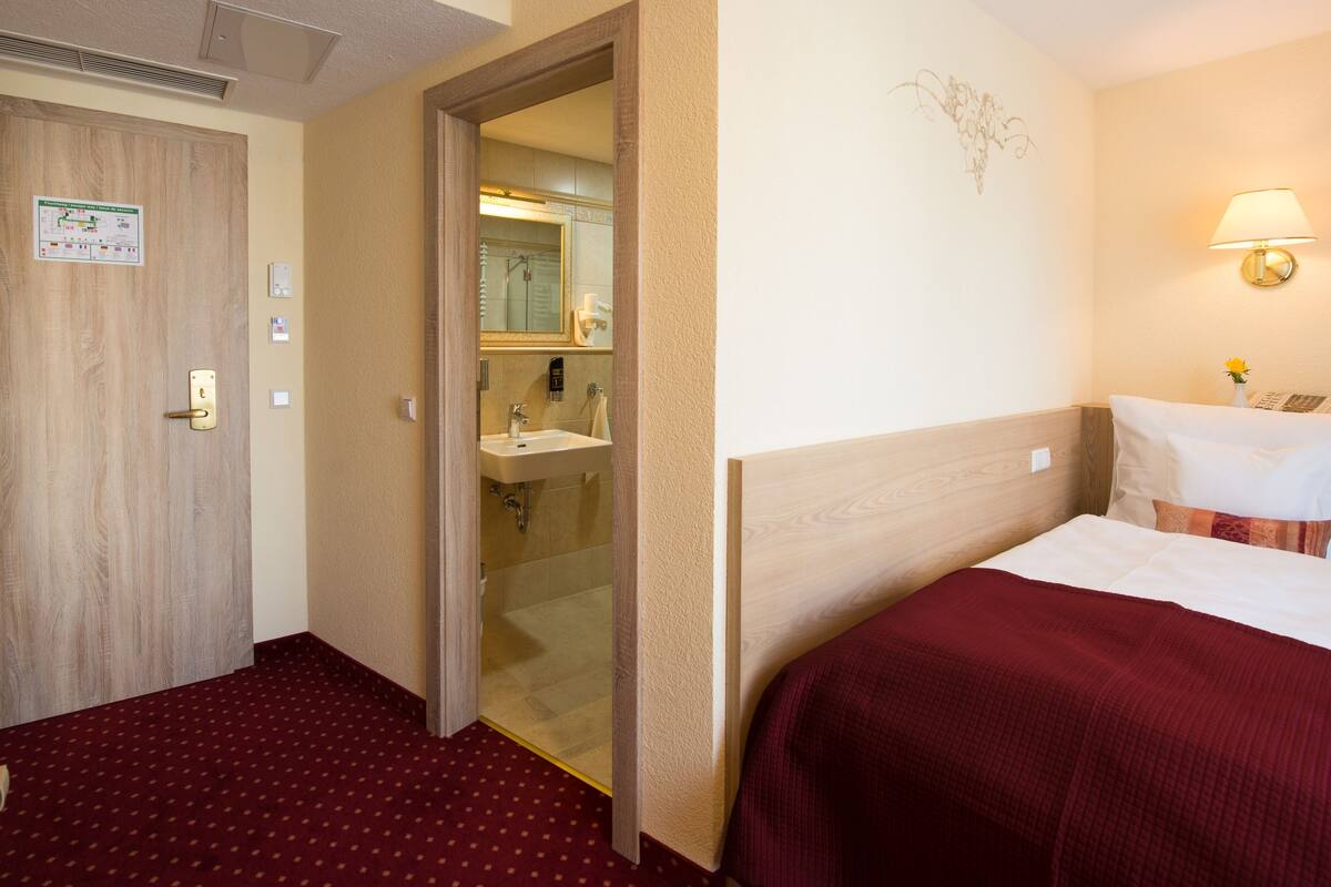 A hotel room with a wooden door, a bathroom with a sink and mirror, a bed with a red blanket, and a wall-mounted lamp.