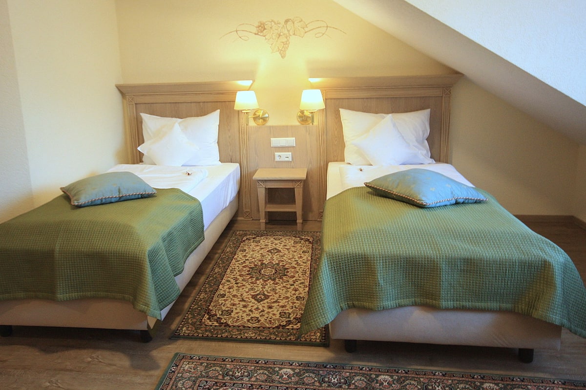 A room with two single beds, each with a green blanket and pillow, a small bedside table, and a decorative wall hanging above the beds.