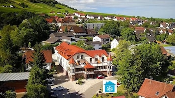 Un edificio de estilo europeo tradicional con techo de tejas rojas, rodeado de exuberante vegetación y un pequeño estacionamiento.