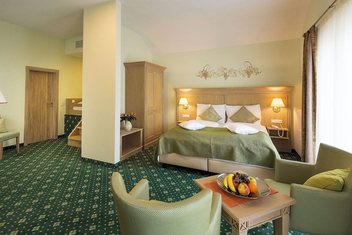 A hotel room with a bed, two chairs, a small table with fruit, a lamp, and a wall decoration.