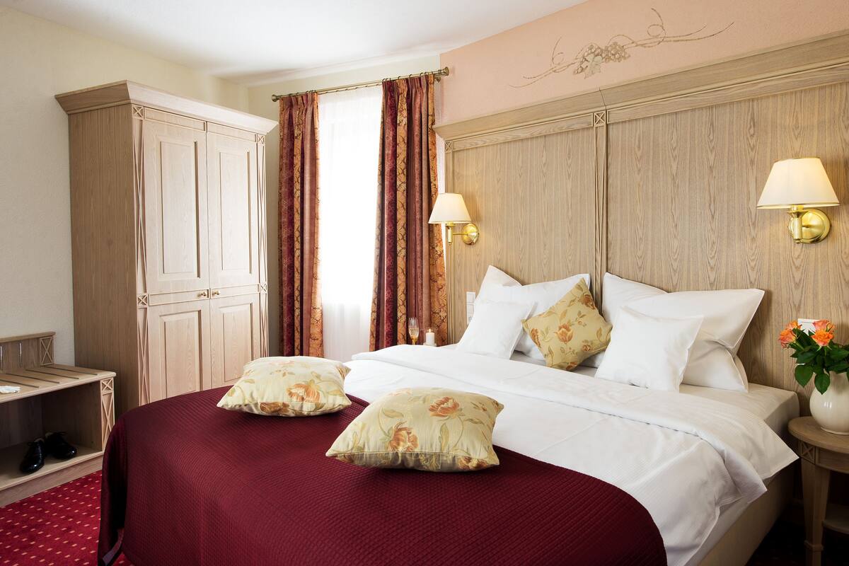 A hotel room with a large bed, two bedside lamps, a wooden headboard, a nightstand with a vase of flowers, and a bench with shoes.