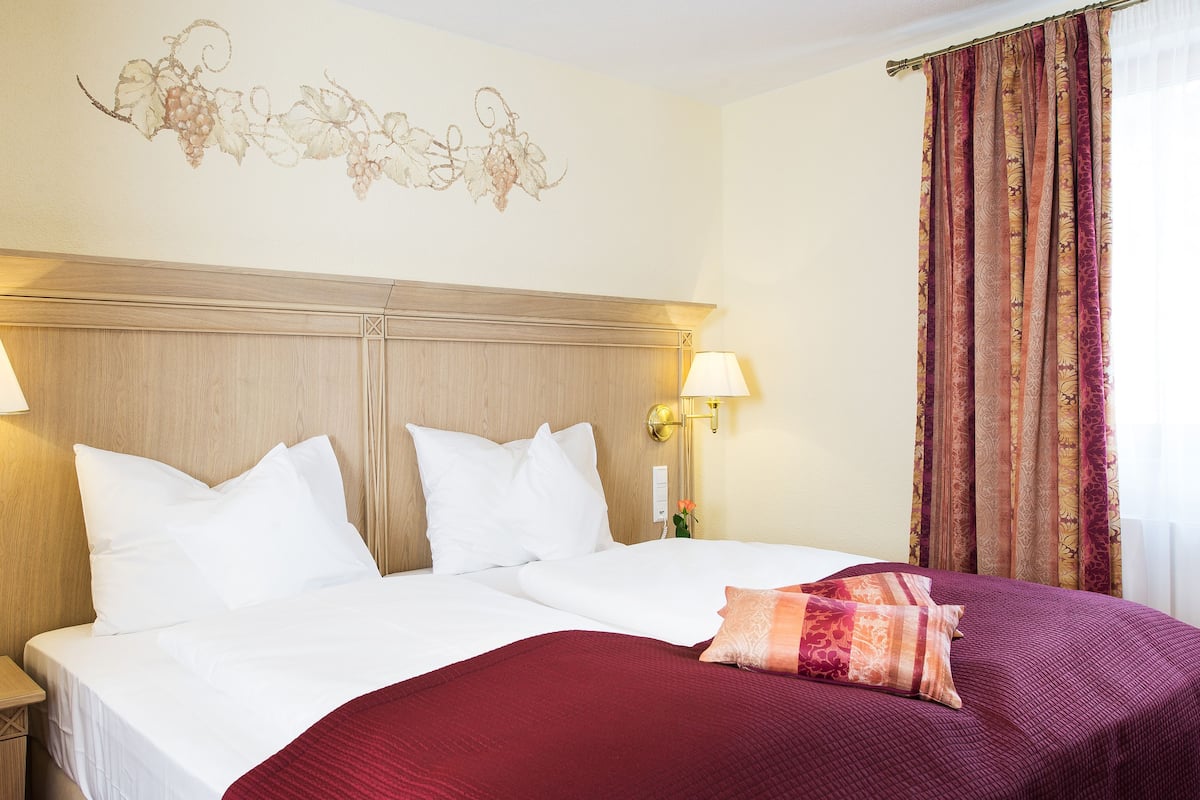 A double bed with white linens, two bedside lamps, a floral wall decoration, and a window with patterned curtains.