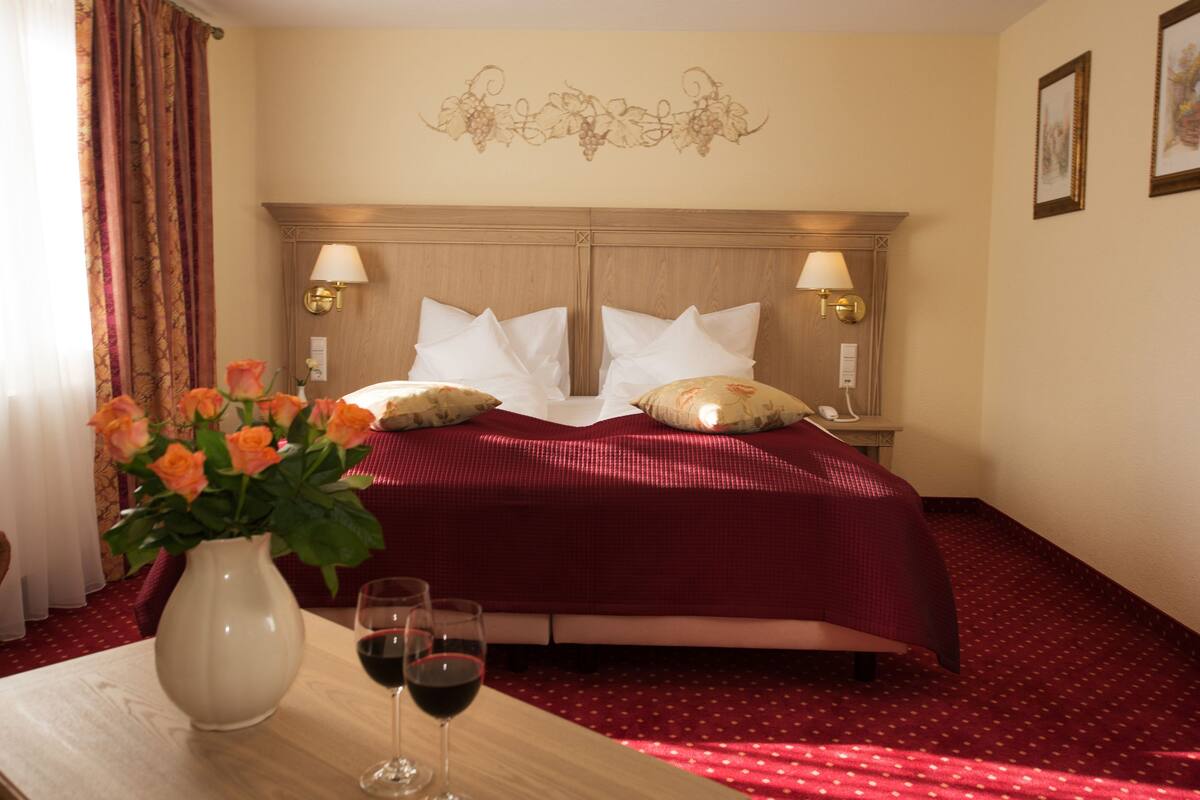 A hotel room with a bed, two bedside lamps, a vase with flowers, and two wine glasses on a table.