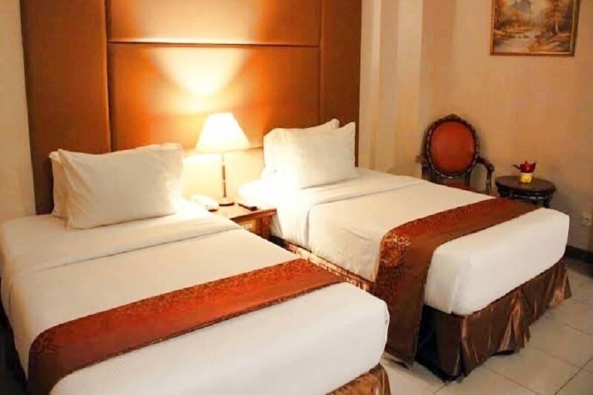 A hotel room with two beds, a nightstand with a lamp, a small table with a vase, and a painting on the wall.
