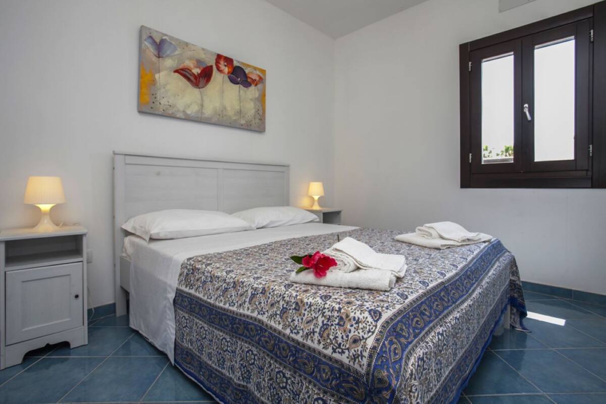 A bedroom with a bed, two bedside lamps, a nightstand, a painting on the wall, and a window with a view.