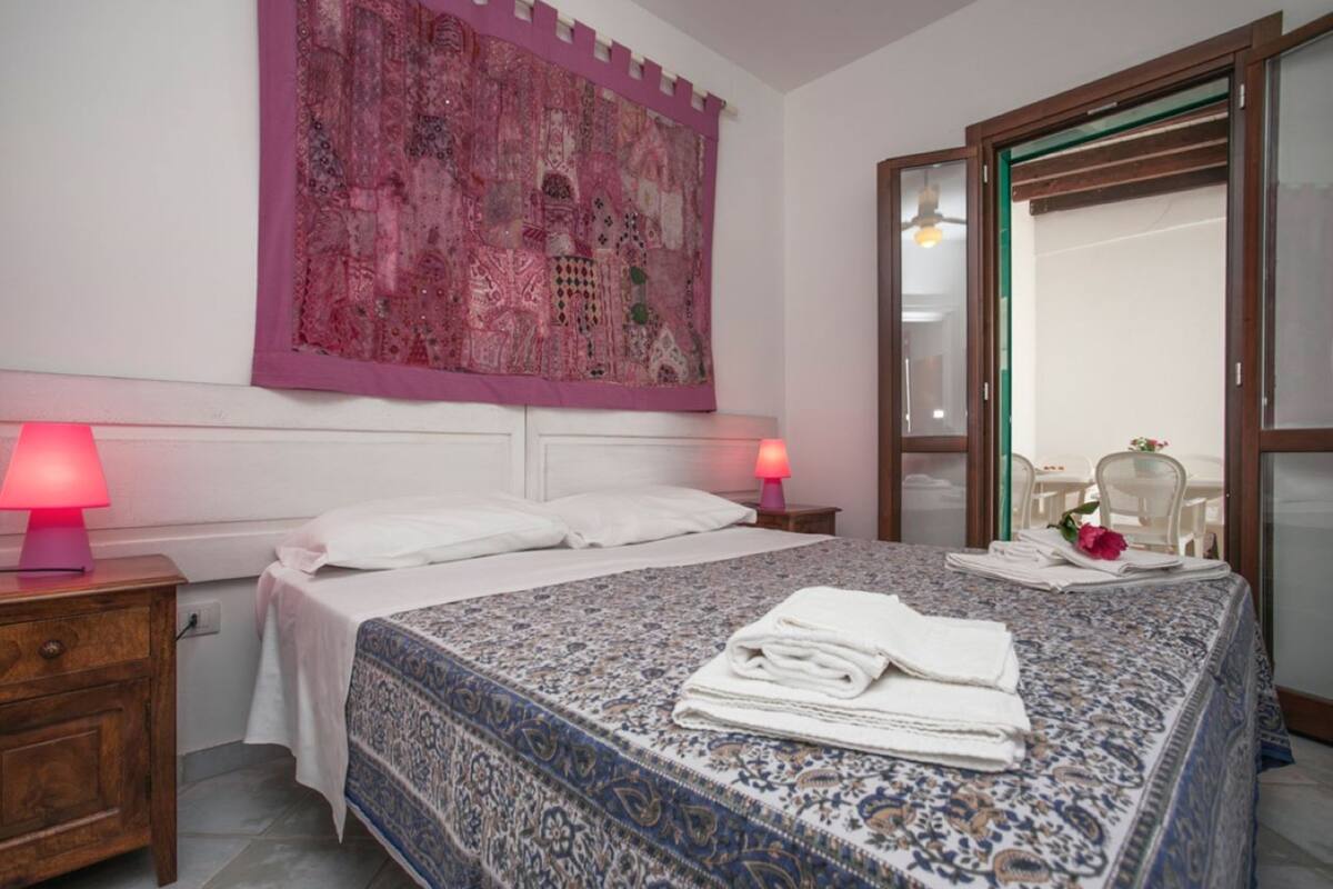 A bedroom with a large bed, a nightstand with a lamp, a window with a view, and a mirror.