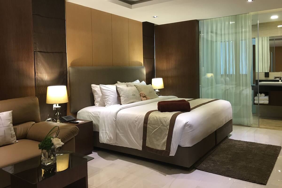 A modern hotel room with a large bed, a sitting area with a sofa, a bedside table with a lamp, and a glass partition wall.