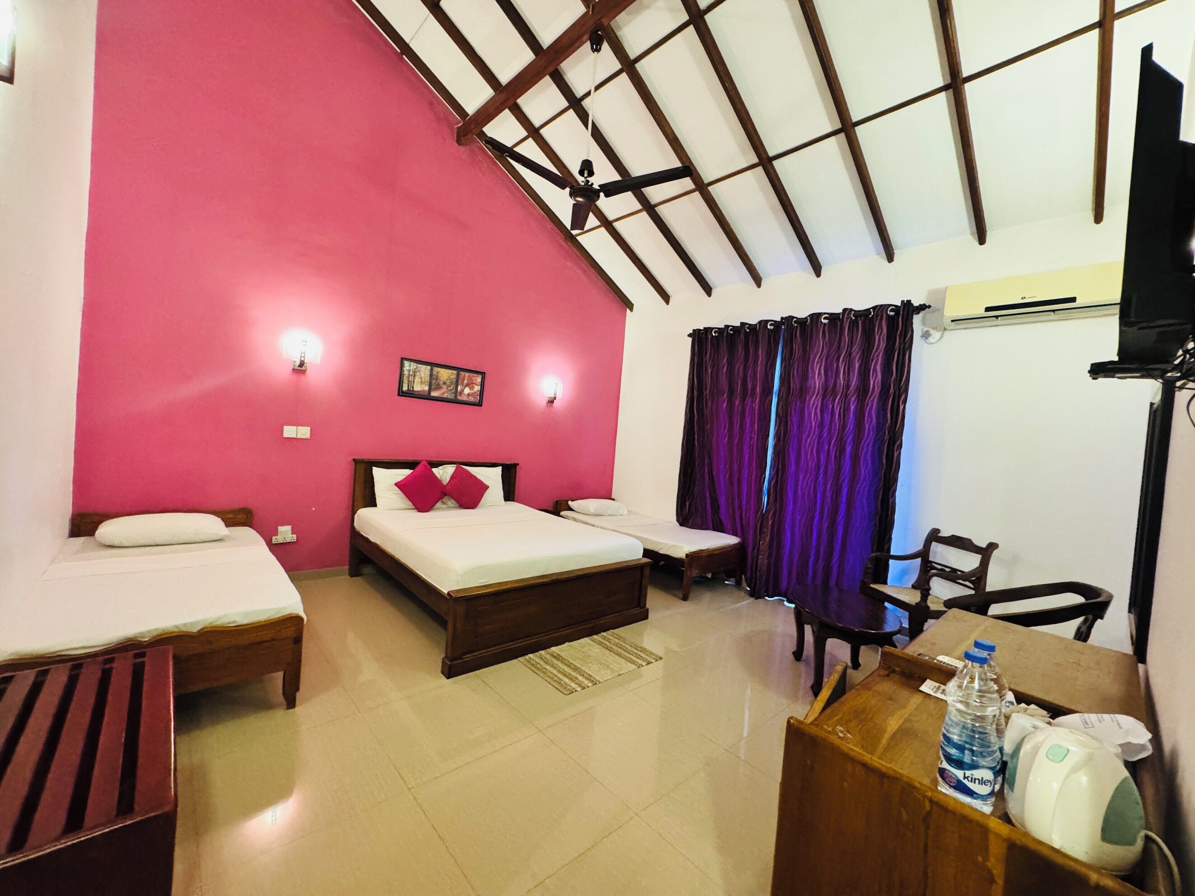 A room with a pink wall, a bed with a wooden frame, a small table, a chair, and a desk with a water bottle and a kettle.