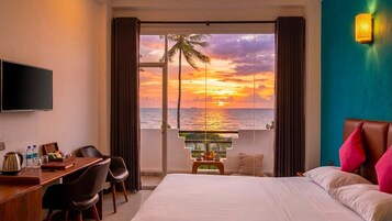 Habitación de hotel con cama, escritorio, silla, televisor y vista al atardecer sobre el océano.