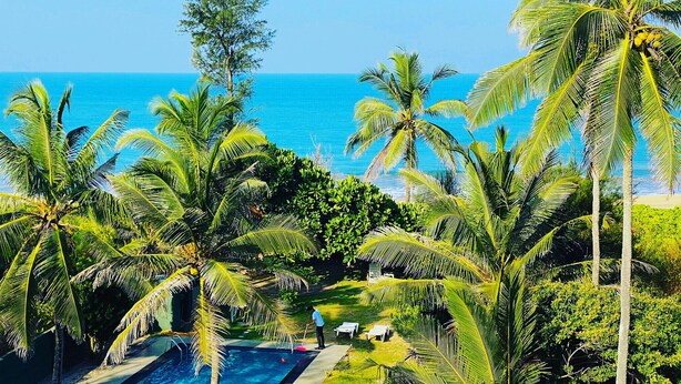 Área de piscina com palmeiras, vista para o oceano e uma pessoa limpando a piscina.