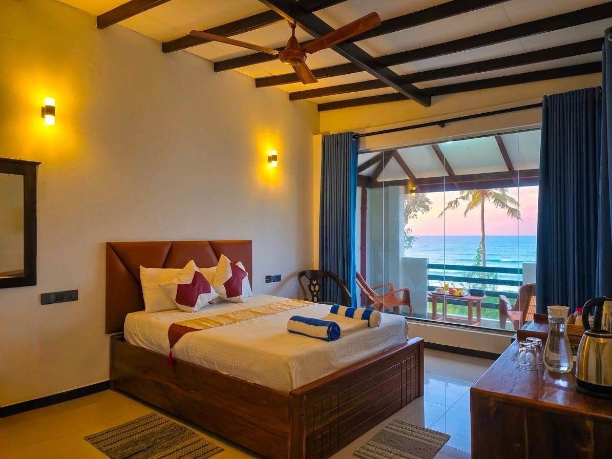 A hotel room with a bed, a ceiling fan, a balcony with a view of the sea, and a table with a pitcher and glasses.