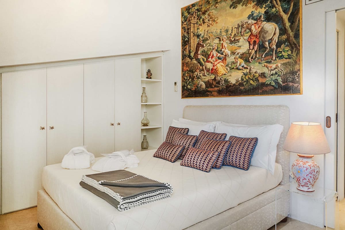 A neatly made bed with a blanket, pillows, and a bedside table with a lamp. There is a painting above the bed and a wardrobe with a mirror.
