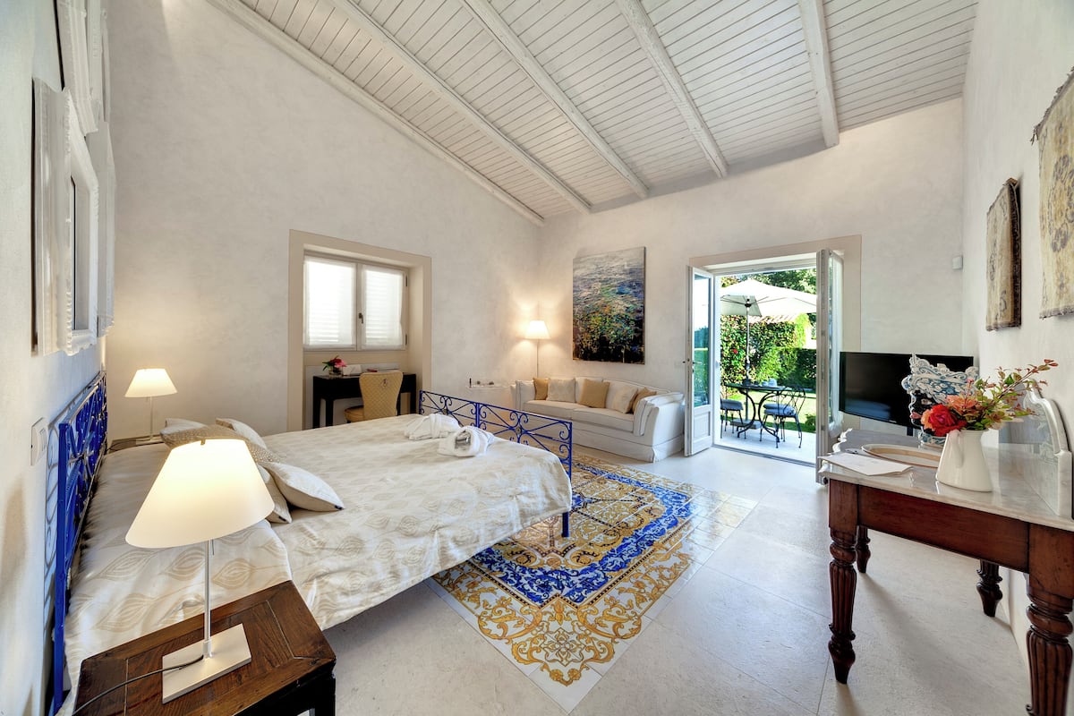 A bedroom with a bed, bedside table, lamp, and a view of the outside.