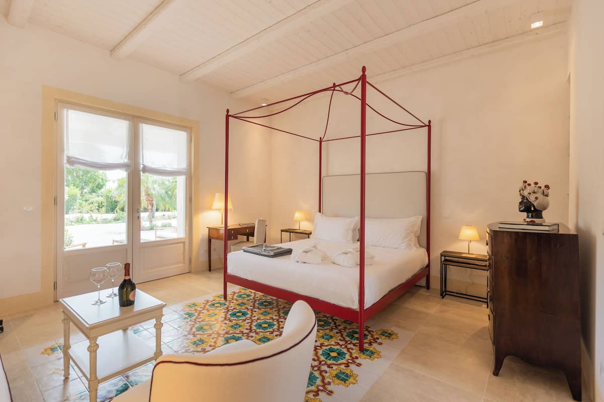 A four-poster bed with a red frame, a white bedspread, and a small table with a bottle and glasses. There is a chair and a nightstand with a lamp.