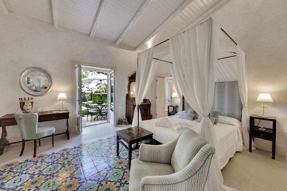A bedroom with a four-poster bed, a sitting area with a chair, a desk, and a small table.