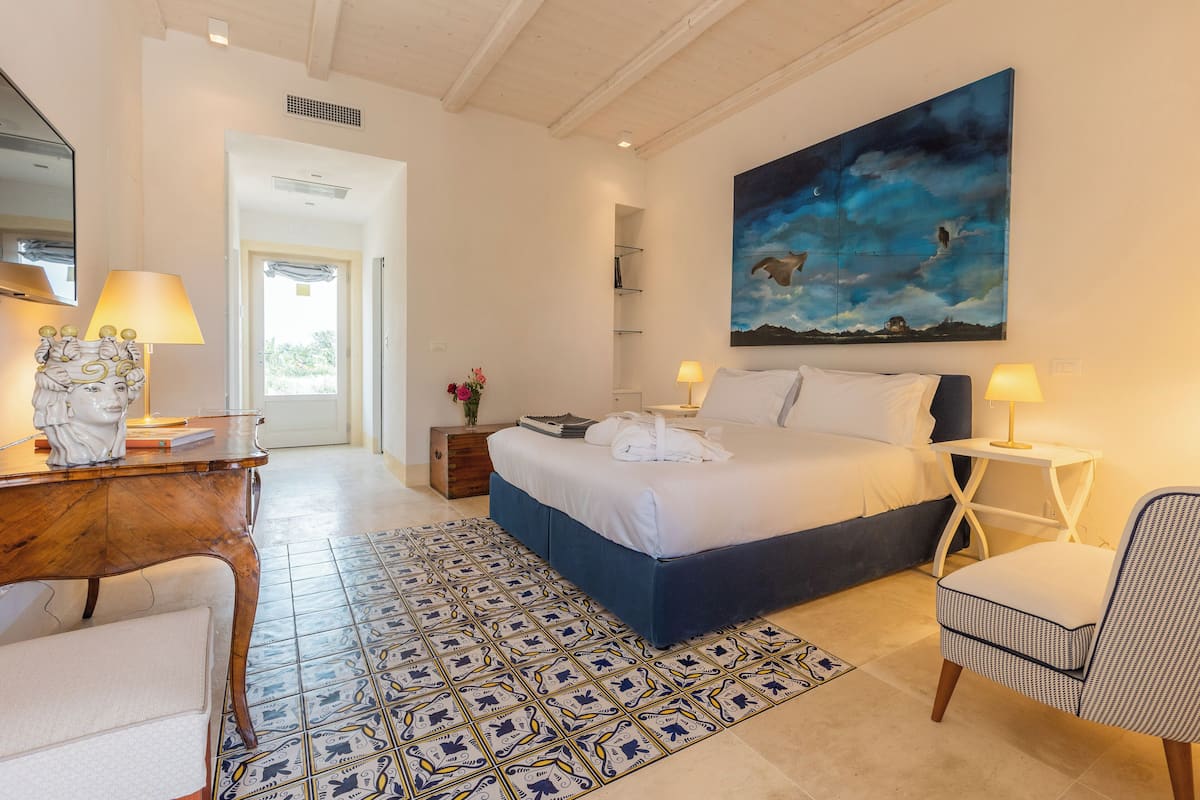A bedroom with a bed, bedside tables, a desk, a chair, a painting, and a patterned rug.
