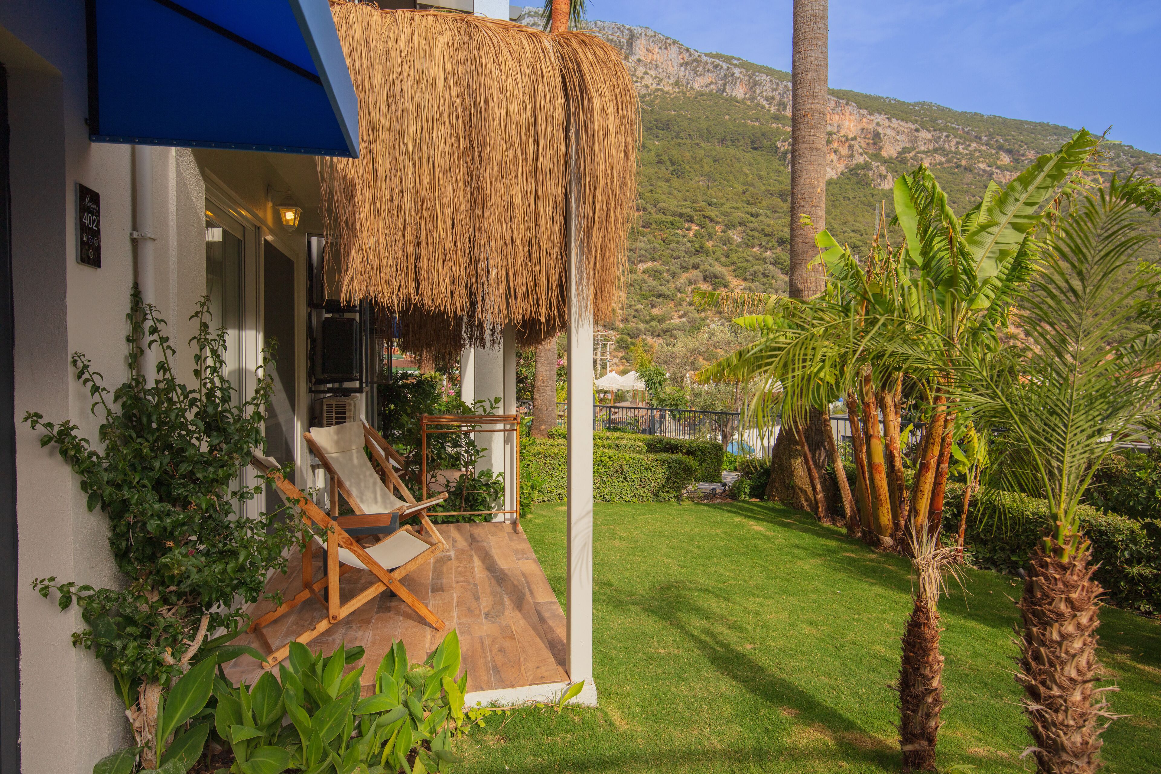 A patio with a thatched roof, a wooden chair, and a grassy lawn with palm trees.