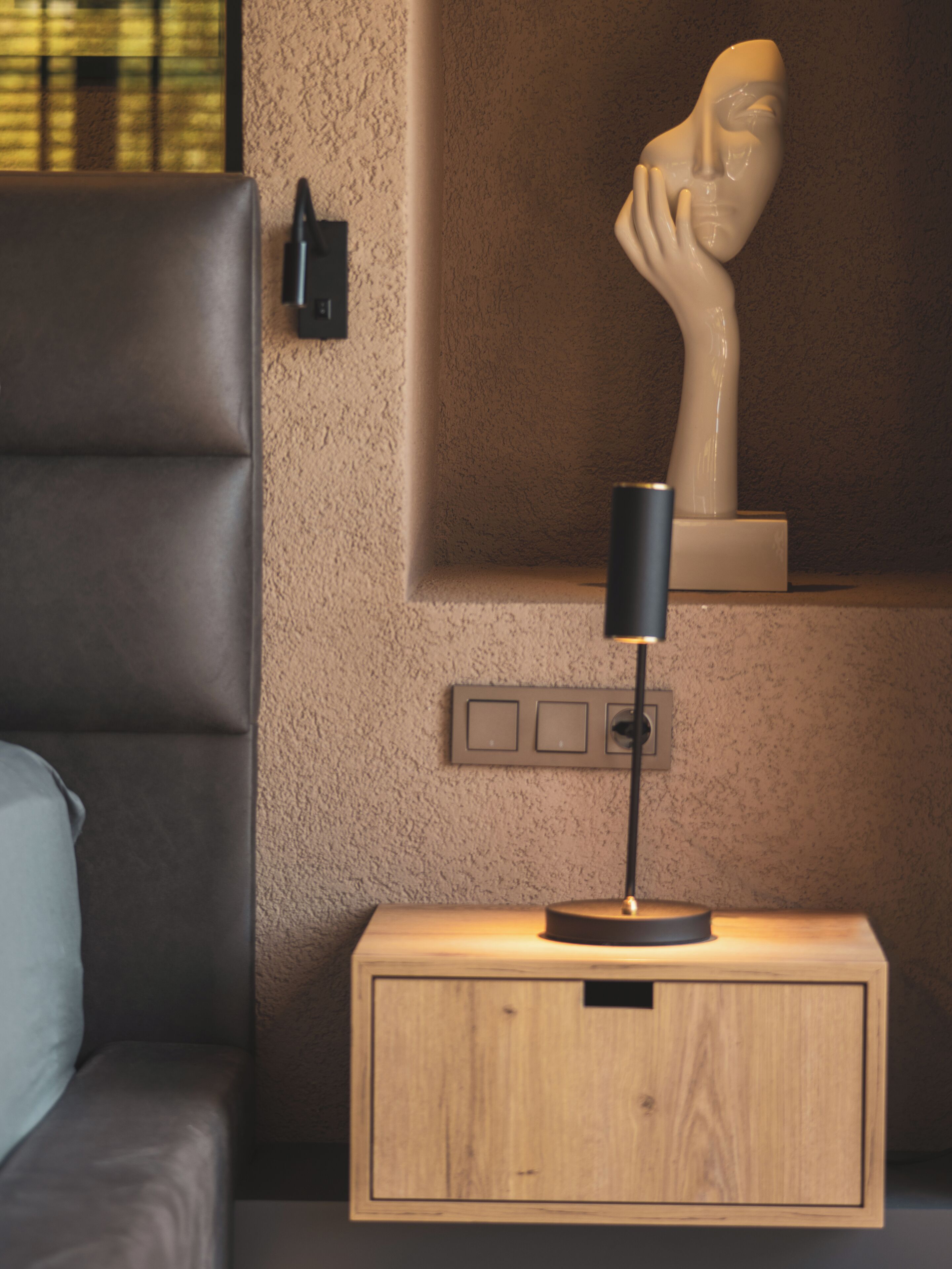 Un dormitorio con una mesita de noche de madera, una lámpara, un busto y un cuadro de interruptores en la pared.