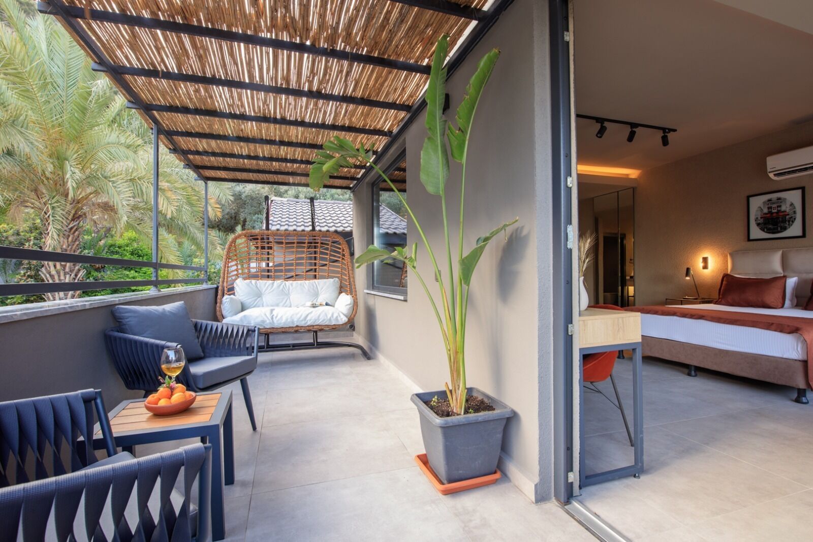 A modern hotel room with a balcony, a wicker sofa, a dining table with a bowl of fruit, and a potted plant.