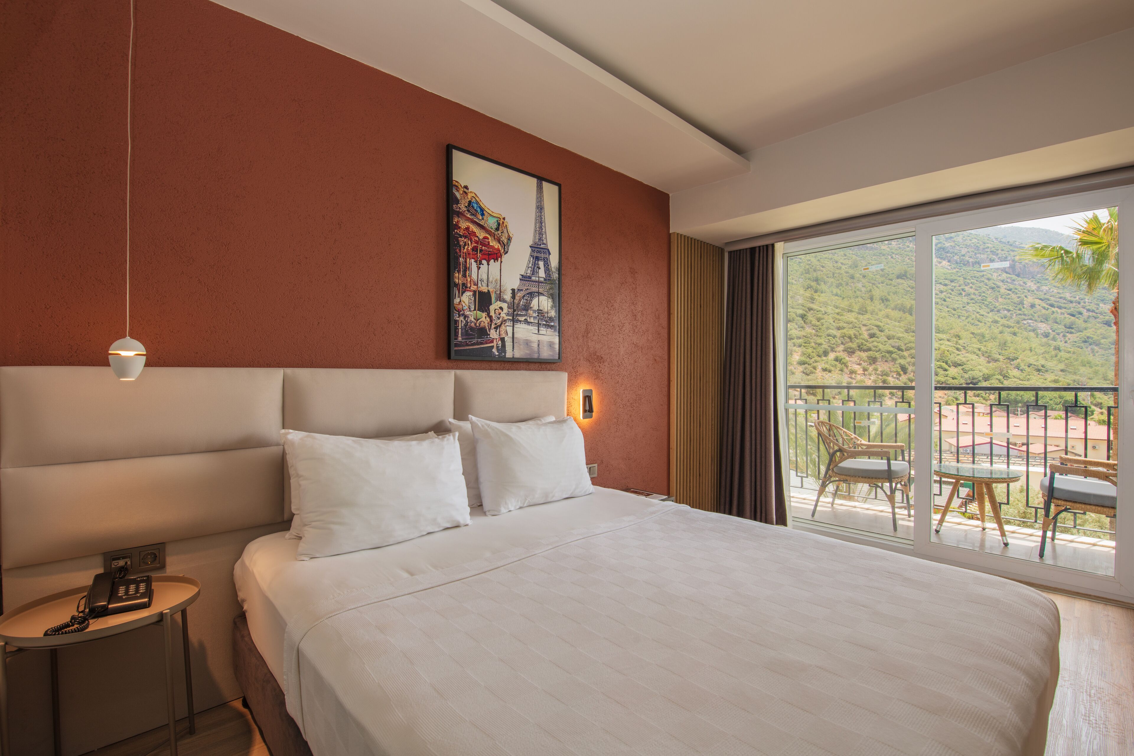 Une chambre d’hôtel avec un grand lit, un tableau représentant la Tour Eiffel, un balcon avec du mobilier d’extérieur et un téléphone sur la table de chevet.