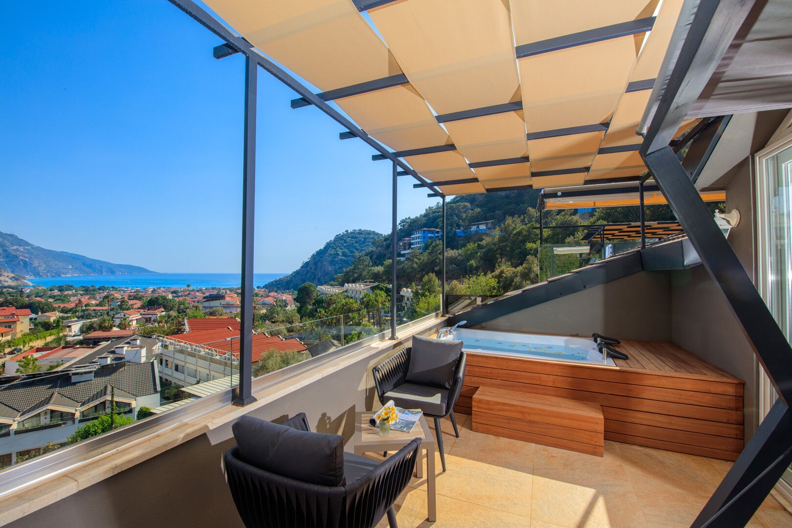 A balcony with a view of a coastal town, a wooden bench, and a table with chairs.
