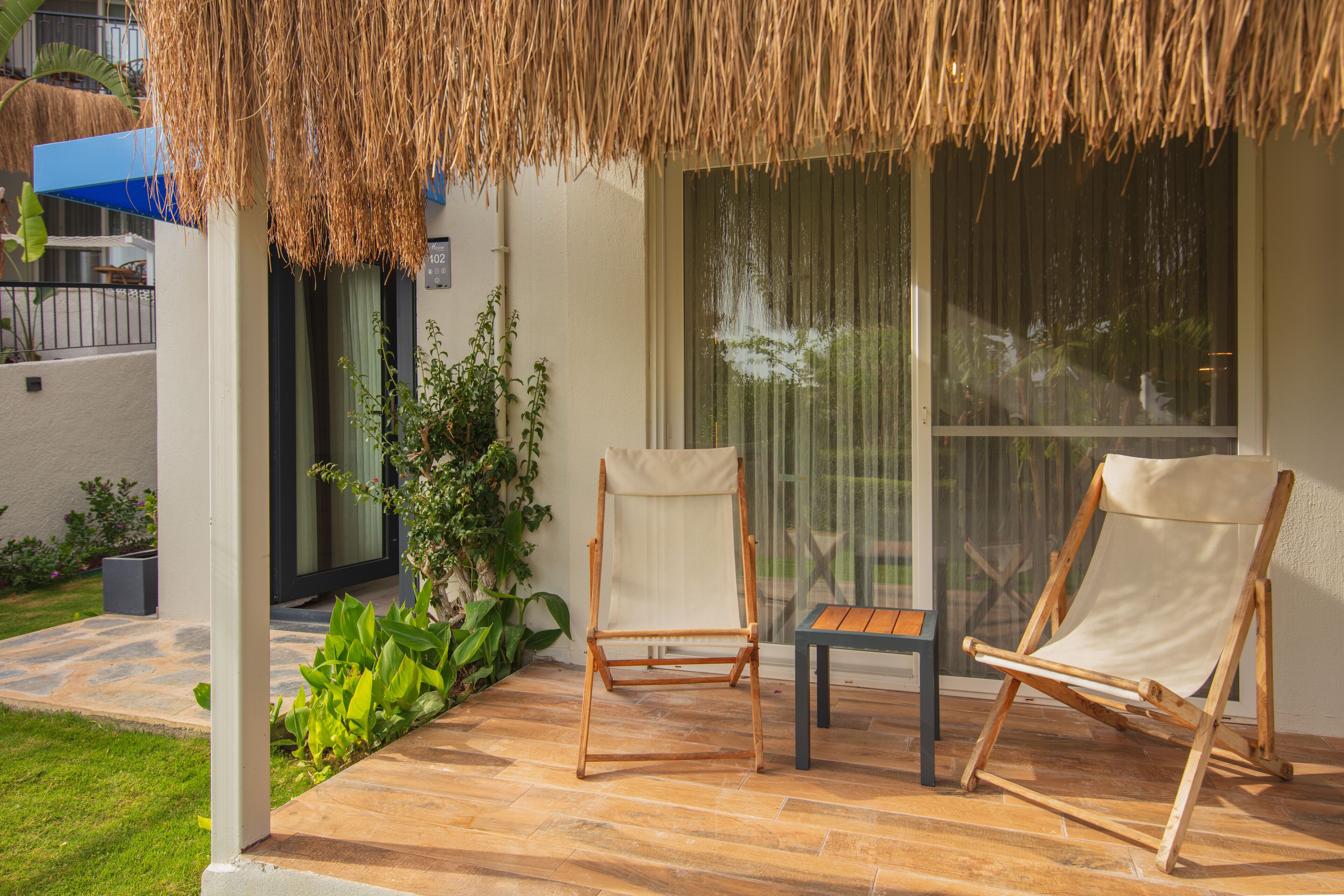 A patio with two wooden chairs and a small table, a thatched roof, and a large window with curtains.