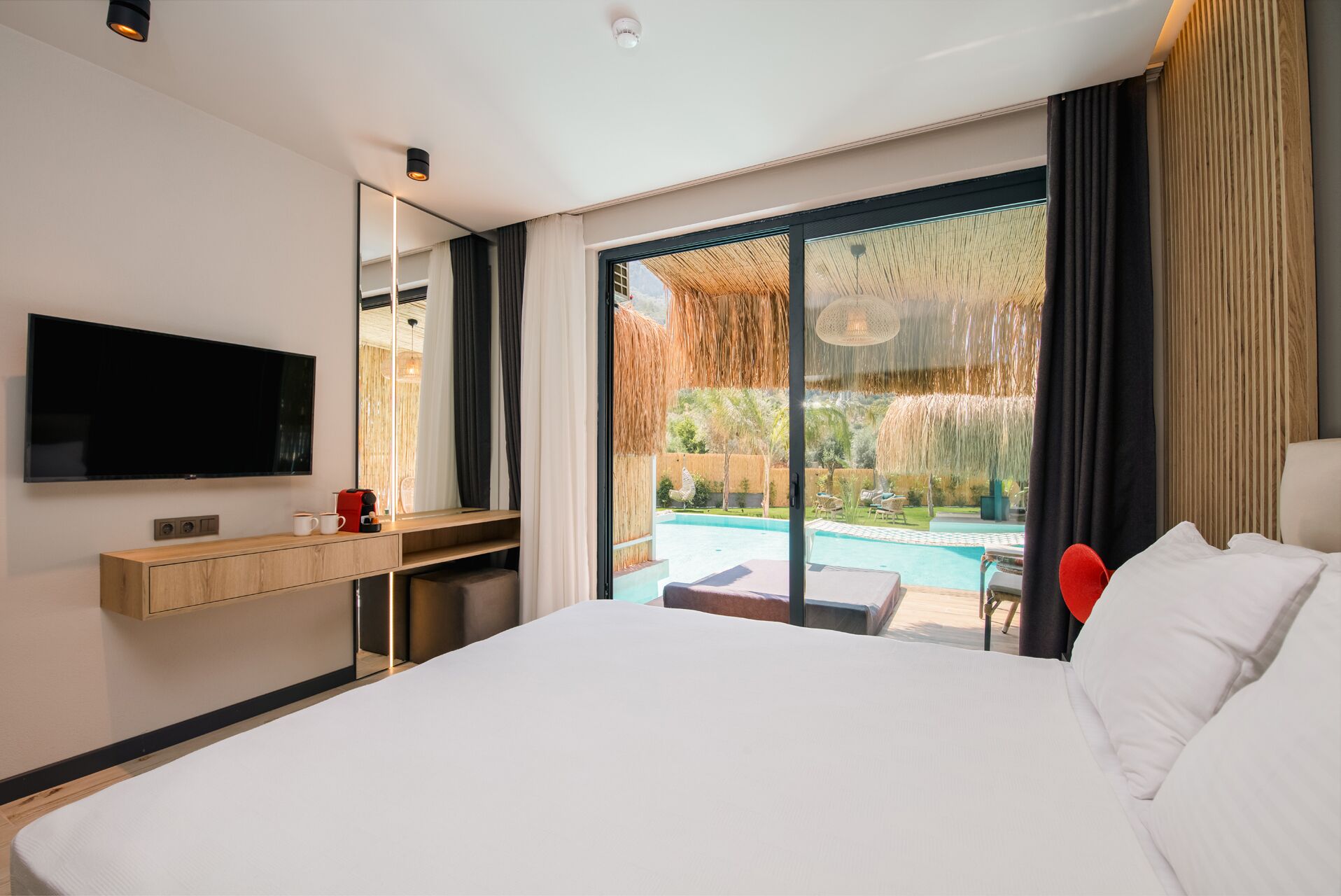 A hotel room with a large bed, a TV mounted on the wall, a wooden desk, and a view of a swimming pool through large glass doors.
