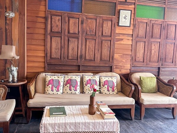 A cozy room with a wooden panel wall, a sofa with elephant-patterned cushions, a side table with a lamp and a decorative elephant figure, and a small table with books and a vase.