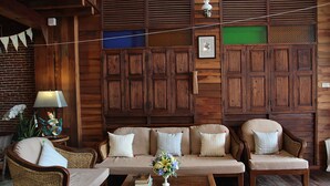 A cozy room with a wooden wall panel, a sofa, two armchairs, a small table with books and a vase, and a lamp. - Rustic River Boutique (Chiang Mai)