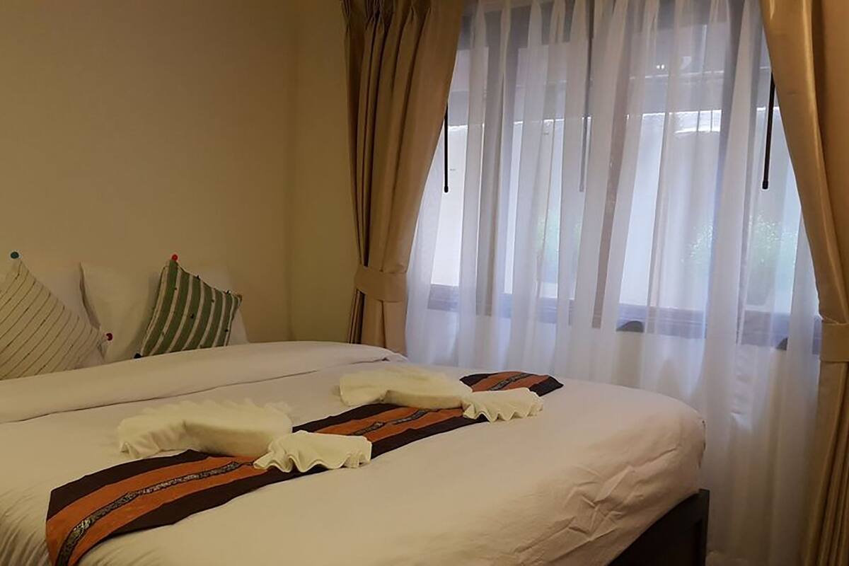 A neatly made bed with white bedding and a striped pillow, a window with sheer curtains, and a plain wall.