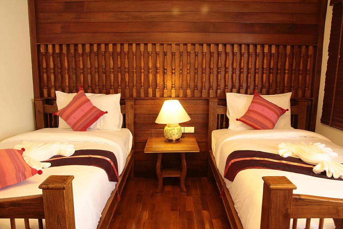 A hotel room with two beds, wooden headboard, a small table with a lamp, and a nightstand with a lamp.