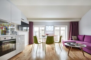 A modern kitchen with white cabinets, a built-in oven, and a coffee maker. There is a dining area with a round table and green chairs. A purple sofa is in the living area, and a coffee table is in front of it.