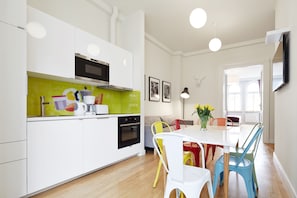 A modern kitchen with white cabinets, a microwave, and a green backsplash. There is a dining area with a white table and colorful chairs. - Crownhill Apartments (Kraków)