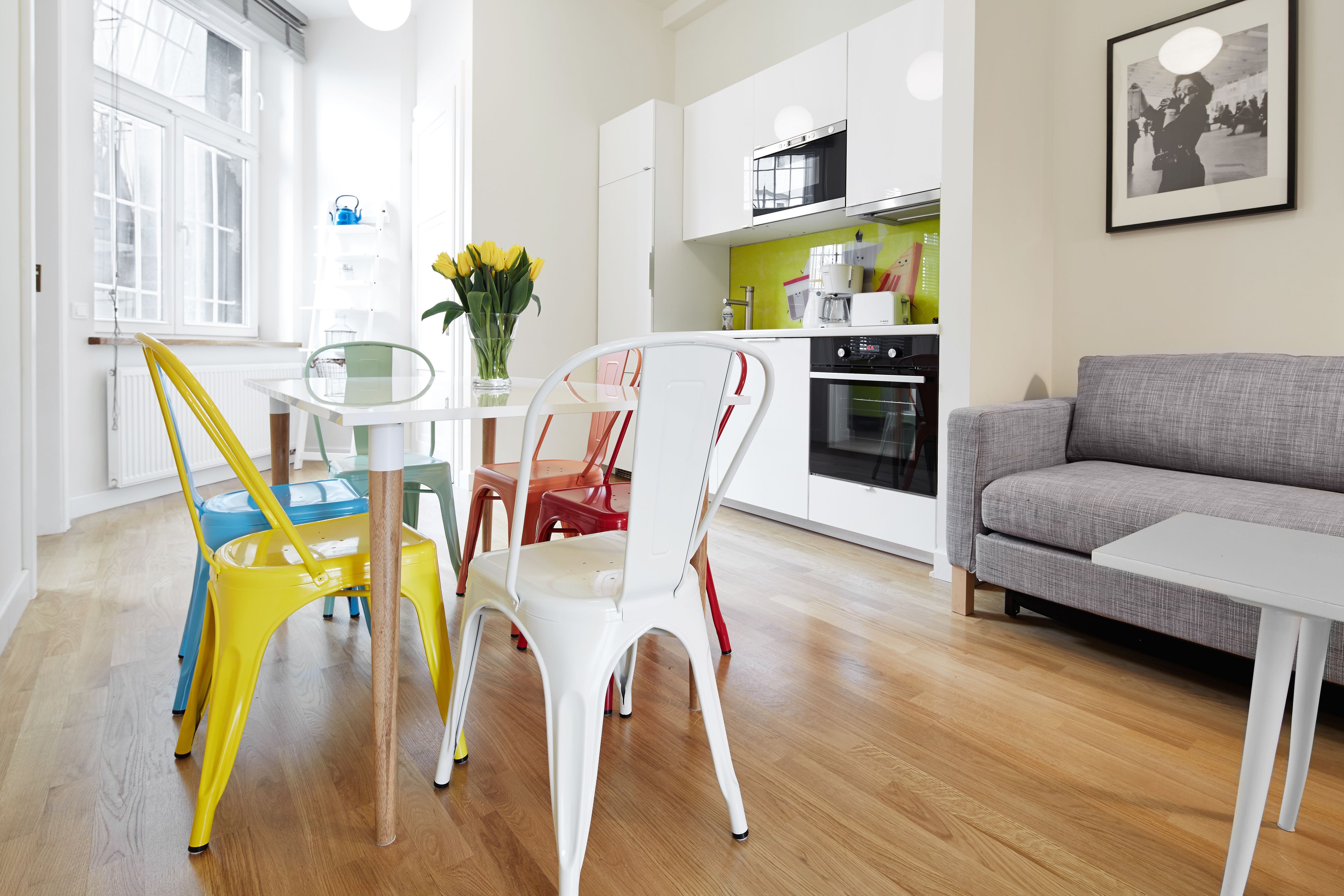 Une cuisine moderne avec des meubles blancs, un canapé gris et des chaises colorées.