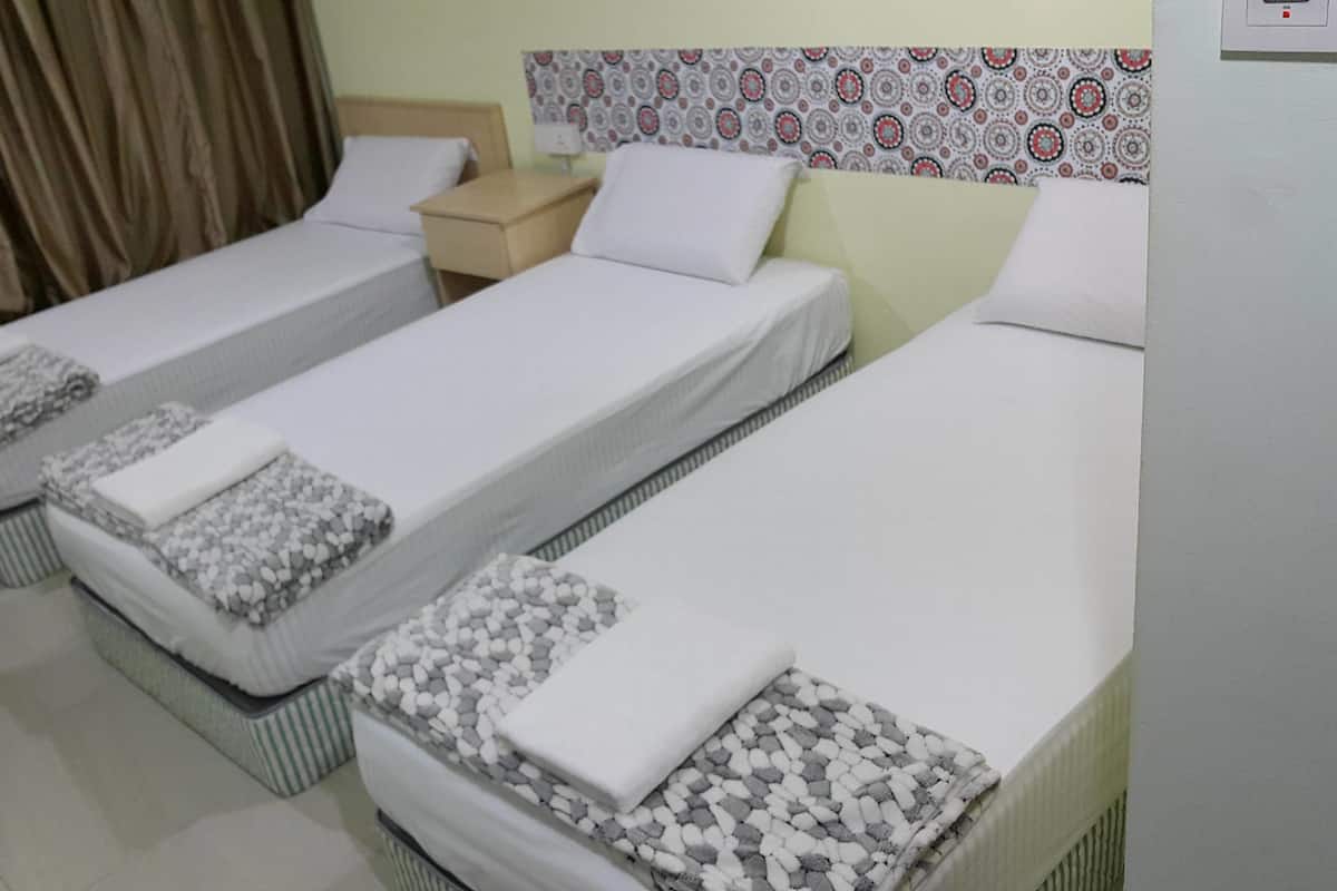 A hotel room with three single beds, each with a pillow and a blanket. There is a nightstand between the beds, and a chair is visible in the corner.