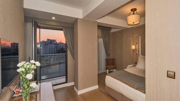 Un cuarto de hotel con una cama, un escritorio con un florero, una silla, una ventana que ofrece vista a un paisaje urbano y una lámpara fijada a la pared.