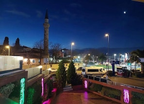 A nighttime view of a cityscape with a prominent tower, a well-lit plaza, and a parking area.