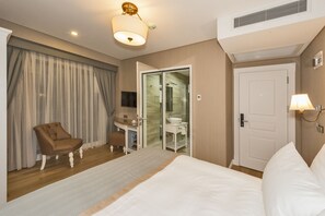 A hotel room with a bed, a chair, a desk with a computer, a bathroom with a glass door, and a door with a light fixture. - Hanna Hotel (Istanbul)