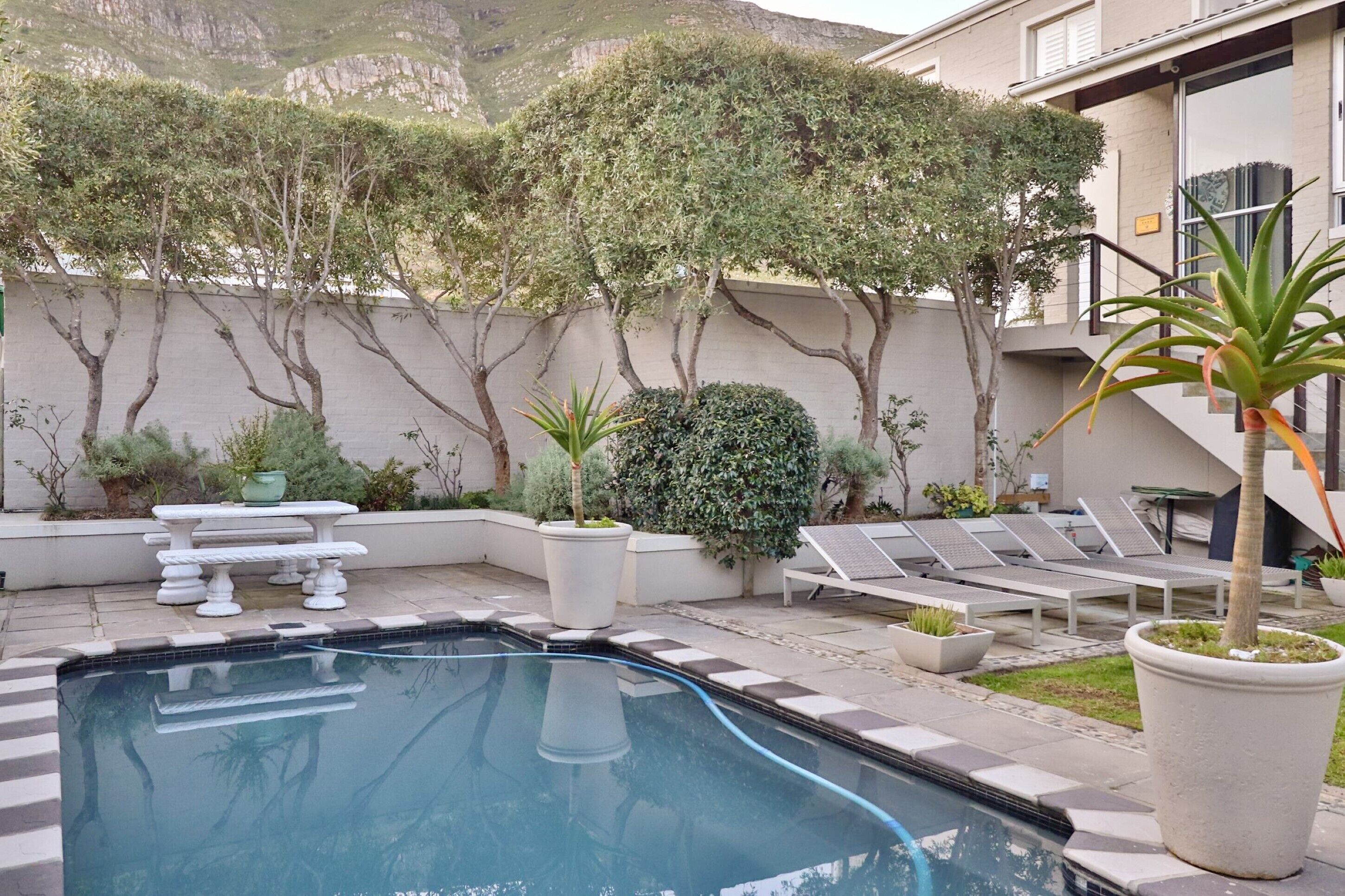 A swimming pool with a tiled deck, surrounded by potted plants, a white table, and lounge chairs.