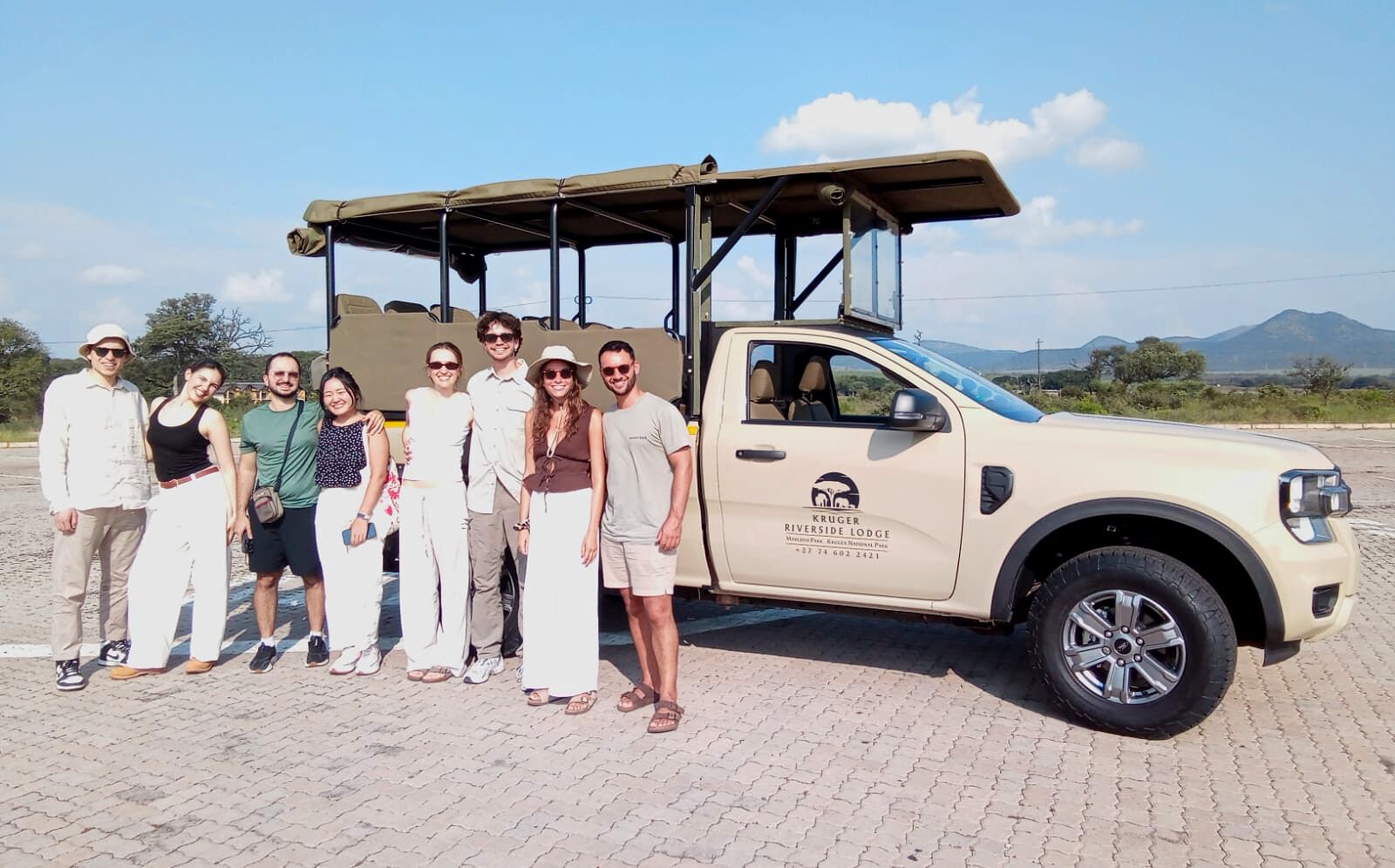 A group of people standing in front of a beige SUV with a canopy, parked on a paved area with a mountainous landscape in the background.